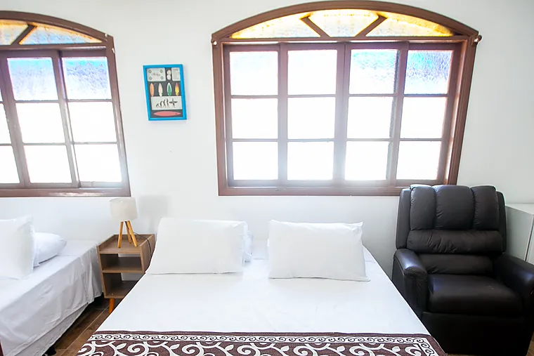 Cama de casal da Suíte Júnior da Petit Refuge entre as janelas arqueadas e a poltrona